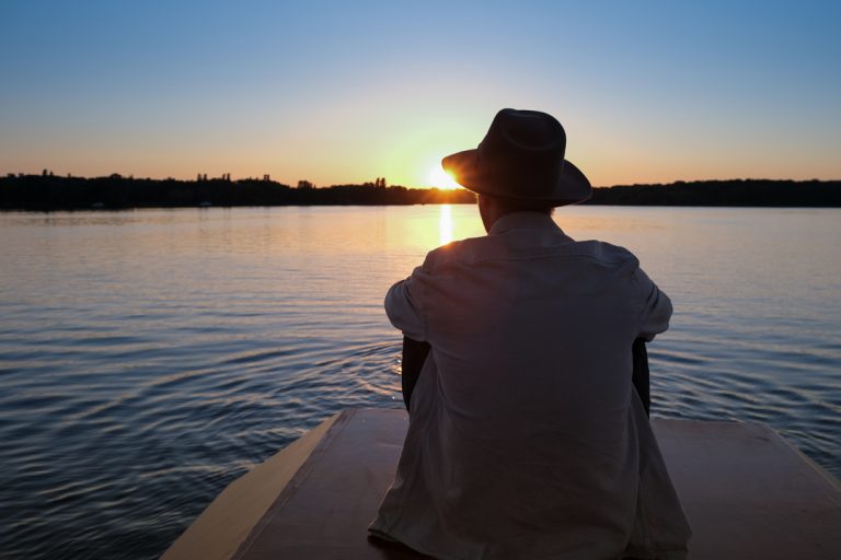 Björn sitzt am Steg und blickt über einen See bei Sonnenuntergang, Moment des Innehaltens beim Entdecken neuer Orte