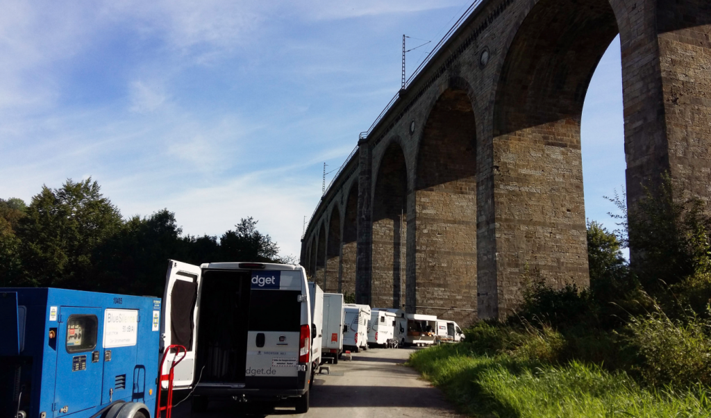 Filmfahrzeuge und Technik an einer abgelegenen Drehbasis unter einer Eisenbahnbrücke