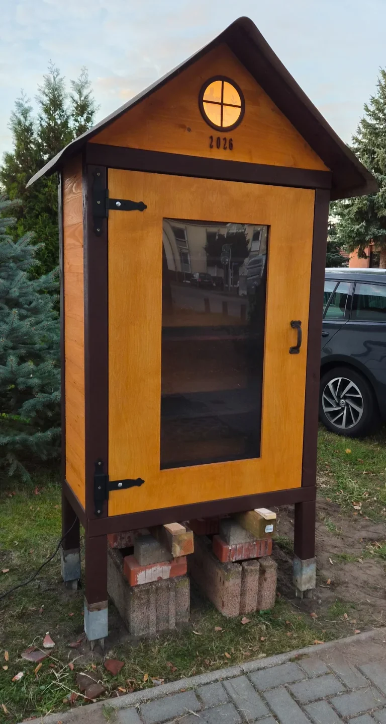 Bücherhaus am Gehweg mit eingebauter Glastür, schwarzen Beschlägen und leuchtendem Giebelfenster; darunter stehen noch provisorische Stützen.